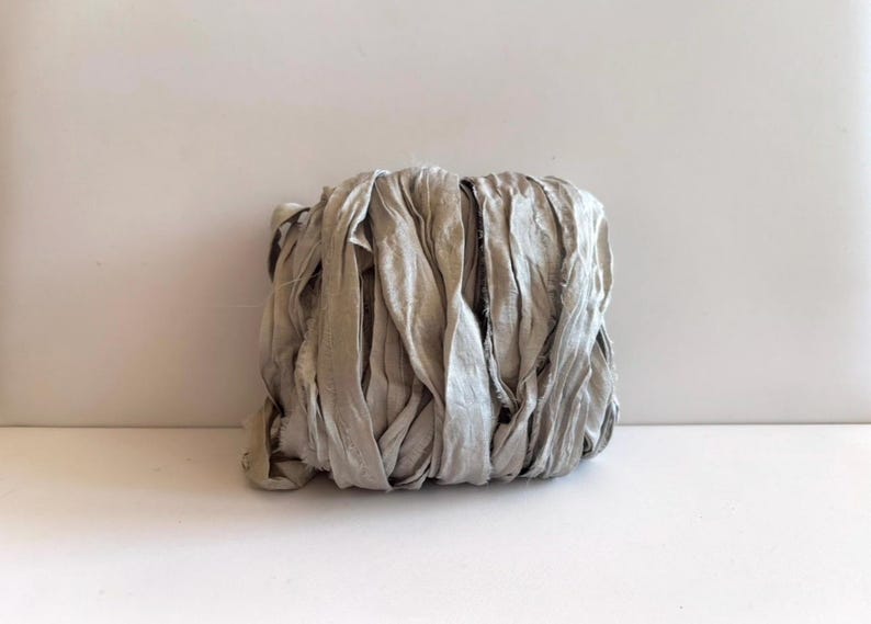May include: A bundle of light gray silk ribbon with a crinkled, textured surface and frayed edges. The ribbon is wound into a compact, rectangular shape, showcasing a soft, matte finish and a consistent neutral color throughout the entire length.