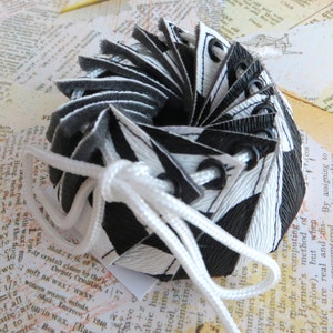 Könnte beinhalten: Eine schwarze und weiße Stoffrosette mit einer weißen Schnur, die um sie herum gebunden ist. Die Rosette besteht aus Stoffstreifen, die zusammengenäht sind, um eine Blütenform zu bilden. Die Rosette ist ein dekoratives Element, das für verschiedene Zwecke verwendet werden kann, z. B. um einem Geschenk oder der Inneneinrichtung einen Hauch von Eleganz zu verleihen.