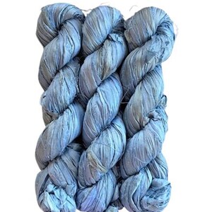 May include: Three skeins of light blue, textured yarn are tightly twisted together. The yarn appears to be made of fabric strips, creating a unique, braided appearance. The color is a soft, muted blue with subtle variations, suggesting a hand-dyed or natural fiber material.
