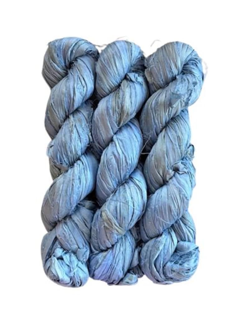 May include: Three skeins of light blue, textured yarn are tightly twisted together. The yarn appears to be made of fabric strips, creating a unique, braided appearance. The color is a soft, muted blue with subtle variations, suggesting a hand-dyed or natural fiber material.
