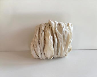 May include: A bundle of off-white silk ribbon featuring a soft, slightly wrinkled texture and frayed edges. The ribbon is coiled into a loose, rounded shape, displaying a subtle sheen and a light, creamy color throughout the entire length.