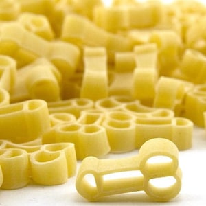 May include: A close-up of a pile of dry pasta shaped like a letter 'T' with a single piece of pasta in the foreground. The pasta is a light yellow color.
