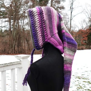 May include: A handmade crochet hood in shades of purple, gray, and pink. The hood features a pointed shape and long, dangling end. The hood is displayed on a black mannequin, with a snowy outdoor background.