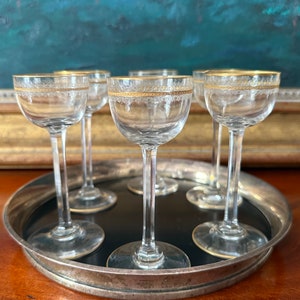 May include: Set of six clear glass cordial glasses with gold rims and delicate etched detailing. The glasses are displayed on a silver-rimmed tray with a black interior, showcasing their elegant design. Perfect for serving after-dinner drinks.