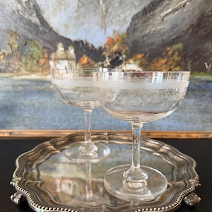 Könnte beinhalten: Zwei klare Champagner-Coupe-Gläser mit geätzten Mustern stehen auf einem silberfarbenen Tablett. Die Gläser haben ein dekoratives Band am Rand. Das Tablett hat einen gewellten Rand und verzierte Füße. Im Hintergrund ist ein Landschaftsgemälde zu sehen.