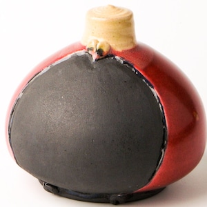 Può includere: Un piccolo vaso rotondo in ceramica con una smaltatura rossa e un'area rotonda nera incassata sul davanti. Il vaso ha un piccolo coperchio rotondo color crema con due piccoli occhi rotondi marroni.