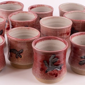 Könnte beinhalten: Set aus zehn roten Keramikbechern mit schwarzen Blattmotiven. Die Becher sind handgefertigt und haben ein rustikales, erdiges Aussehen.