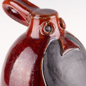 Może przedstawiać: Ceramiczny ptak w kolorze czerwonym z czarnym dziobem i oczami. Ptak ma uchwyt na górze głowy i czarną plamę na brzuchu.