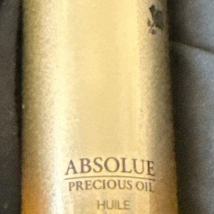Puede incluir: Una botella dorada de Aceite Precioso Absolue de Lancôme. La botella tiene un tapón cuentagotas y está etiquetada con el nombre y la descripción del producto. El aceite es para nutrir e iluminar la piel.