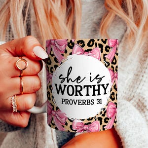 Peut inclure: Mug en c&eacute;ramique blanche avec un motif de n&oelig;uds roses, un fond l&eacute;opard et le texte "she is WORTHY PROVERBS 31". Le mug est tenu par une personne aux longs cheveux blonds et portant des bagues.