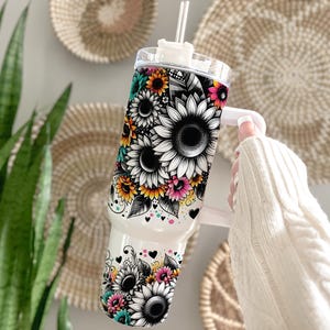 May include: White stainless steel tumbler with a black, white, and yellow sunflower design. The tumbler has a lid and a handle.