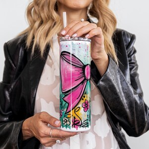 May include: A white tumbler with a pink bow design and floral accents. The tumbler has a straw and a lid. The design includes a large pink bow, flowers, and green and yellow brushstrokes. The tumbler is held by a person.