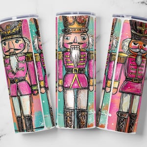 Op de afbeelding: Drie hoge, cilindrische bekers met een kleurrijk, grillig ontwerp met een notenkrakersoldaat. De notenkraker is afgebeeld in roze, goud en zwart, met een kroon en een lange witte baard. De achtergrond heeft roze en turquoise penseelstreken.