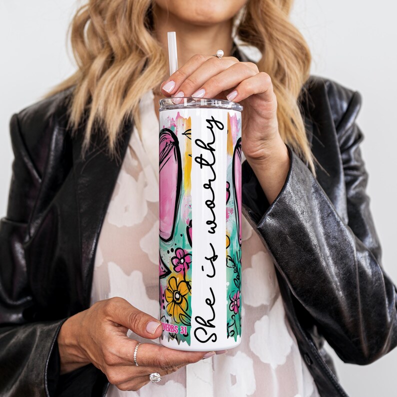May include: A white stainless steel tumbler with a colorful floral design and the text "She is worthy" in black script. The tumbler features a pink and green floral pattern with a straw.
