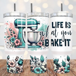 Puede incluir: Un vaso blanco con un gráfico de batidora de pie de color verde azulado y plateado y un diseño floral. El vaso tiene una tapa y una pajita. El texto "Life is what you bake it" está impreso en el vaso.