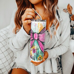 May include: A tall, stainless steel tumbler with a colorful design featuring a large pink bow, flowers, and paint splatters. The tumbler has a silver lid and a metal straw. The overall design is bright and cheerful.