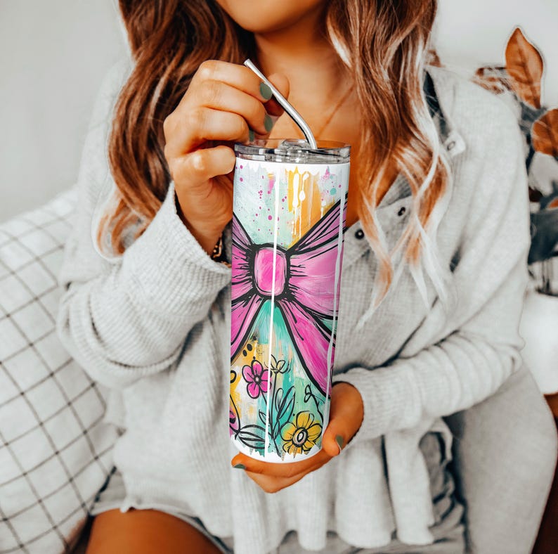 May include: A tall, stainless steel tumbler with a colorful design featuring a large pink bow, flowers, and paint splatters. The tumbler has a silver lid and a metal straw. The overall design is bright and cheerful.