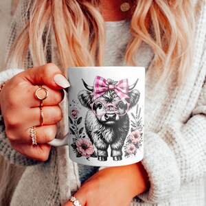 Peut inclure: Mug en c&eacute;ramique blanche avec une illustration en noir et blanc d'une vache &eacute;cossaise portant des lunettes roses et un n&oelig;ud en vichy rose et blanc. La vache est entour&eacute;e de fleurs roses.