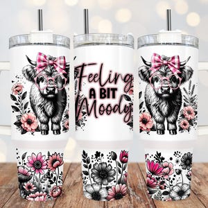 May include: White stainless steel tumbler with a pink and black Highland cow design. The tumbler has a black lid with a straw and says "Feeling a bit Moody".