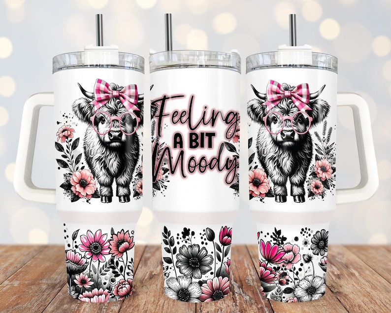 May include: White stainless steel tumbler with a pink and black Highland cow design. The tumbler has a black lid with a straw and says "Feeling a bit Moody".