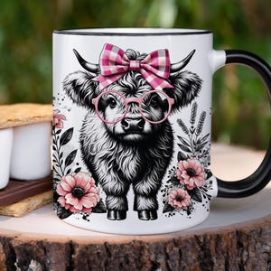 Peut inclure: Une tasse en c&eacute;ramique blanche avec une anse noire. La tasse pr&eacute;sente une illustration en noir et blanc d'une vache des Highlands portant des lunettes roses et un n&oelig;ud en vichy rose et blanc. La vache est entour&eacute;e de fleurs roses et de feuilles vertes.
