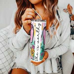 May include: A stainless steel tumbler with a colorful floral and bunny ear design. The tumbler has the words "She is worthy" in a vertical script. The tumbler is held by a person with a silver straw.