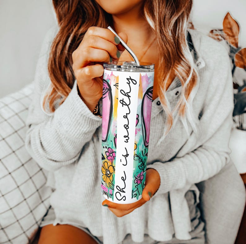May include: A stainless steel tumbler with a colorful floral and bunny ear design. The tumbler has the words "She is worthy" in a vertical script. The tumbler is held by a person with a silver straw.