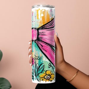 May include: A tall, stainless steel tumbler with a colorful abstract design. The design features a large pink bow with black outlines, yellow and teal brushstrokes, and floral accents. The tumbler is held by a person.