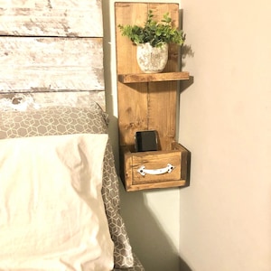 May include: Rustic wooden bedside shelf with a small drawer, a top shelf, and a white metal handle. A small potted plant sits on the top shelf.