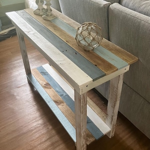 May include: A rustic wooden console table with a distressed white finish. The table top is painted in shades of blue, white, and brown, creating a striped pattern. The table has a lower shelf and is perfect for a coastal-themed living room.