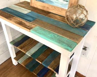 May include: A white wooden side table with three shelves. The shelves are made of reclaimed wood painted in shades of blue, green, and brown. A framed painting of a wave is on the top shelf, and a glass buoy is on the right side of the top shelf.
