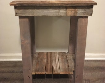 May include: A rustic wooden end table with a single shelf. The table is painted a light gray color and has a distressed finish. The table top is made of reclaimed wood planks. A vintage alarm clock and a framed sign with the number 8 and the word "eight" are on top of the table.