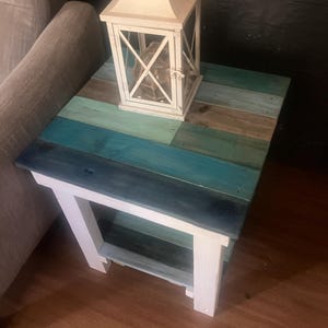 May include: A square side table with a multi-colored plank top featuring shades of blue, teal, and brown, supported by white wooden legs and a lower shelf. A white metal lantern with a glass pane sits on the surface of the table.