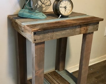 May include: A rustic wooden side table with a distressed finish. The table has a top shelf and a lower shelf, both made of reclaimed wood planks in shades of brown, blue, and gray. A vintage metal fan and an alarm clock are on the top shelf.