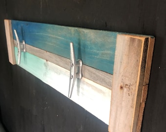 May include: A wooden wall-mounted coat rack with two silver boat cleat hooks. The rack is made of reclaimed wood painted in shades of blue, gray, and white. The wood has a rustic, weathered appearance, adding to its nautical theme.