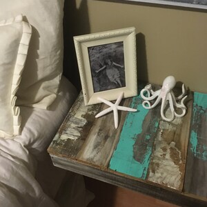 May include: A rustic wooden nightstand with a turquoise blue paint finish. The nightstand has a white picture frame with a black and white photo of a girl in a dress. There is a white starfish and a white octopus figurine on the nightstand.