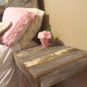 May include: A rustic wooden bedside table with a whitewashed finish. The table is attached to the wall and has a small pink flower pot on top.