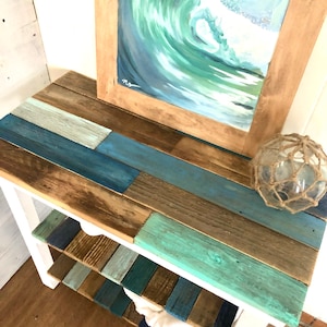 May include: A white wooden shelf unit with three shelves. The shelves are made of reclaimed wood painted in shades of blue, green, and brown. A framed painting of a blue and green wave is on top of the shelf unit. A decorative net ball is on the top shelf.