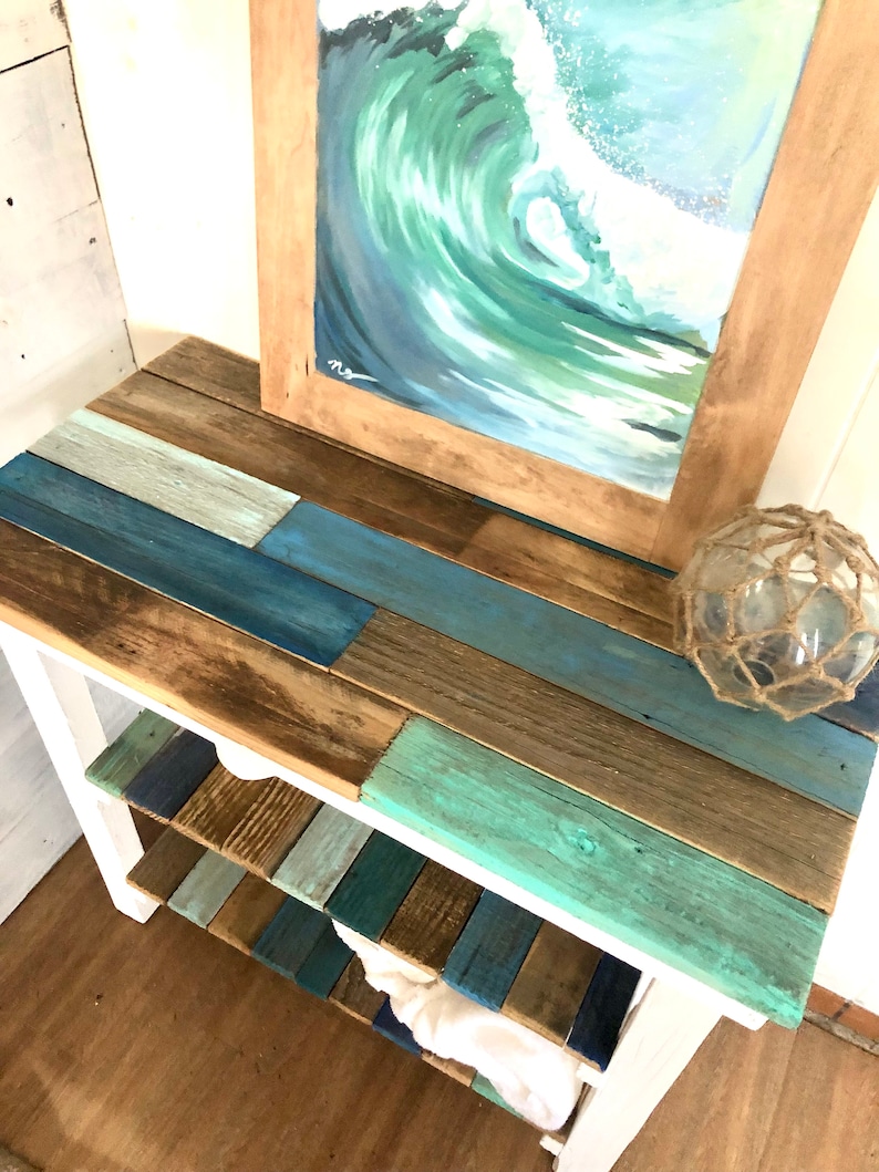 May include: A white wooden shelf unit with three shelves. The shelves are made of reclaimed wood painted in shades of blue, green, and brown. A framed painting of a blue and green wave is on top of the shelf unit. A decorative net ball is on the top shelf.