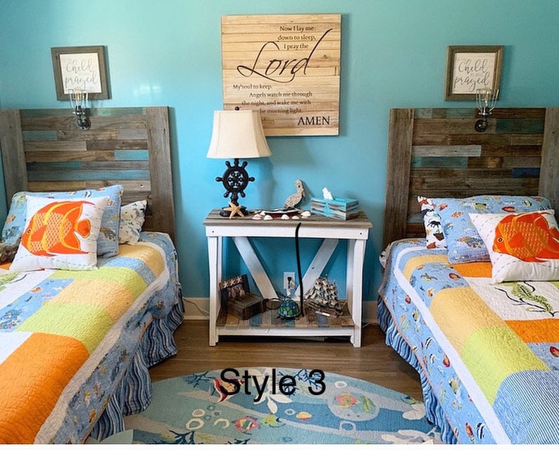 May include: A nautical-themed bedroom with two twin beds, each with a colorful quilt and fish-shaped pillows. A wooden sign with text hangs above a nightstand with a lamp and ship wheel design. The room has a blue wall and a rug with sea creatures.