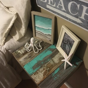 May include: A rustic wooden side table with a turquoise blue paint finish. The table has a white ceramic octopus figurine, a white starfish, and two framed pictures on top. One picture is a seascape painting and the other is a black and white photo. The wall behind the table has a sign that says "BEACH" with an arrow pointing to the right.