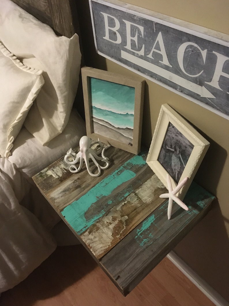 May include: A rustic wooden side table with a turquoise blue paint finish. The table has a white ceramic octopus figurine, a white starfish, and two framed pictures on top. One picture is a seascape painting and the other is a black and white photo. The wall behind the table has a sign that says "BEACH" with an arrow pointing to the right.