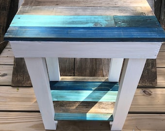 May include: A small white wooden end table with a blue and brown striped top and a lower shelf. The table is sitting on a wooden deck.