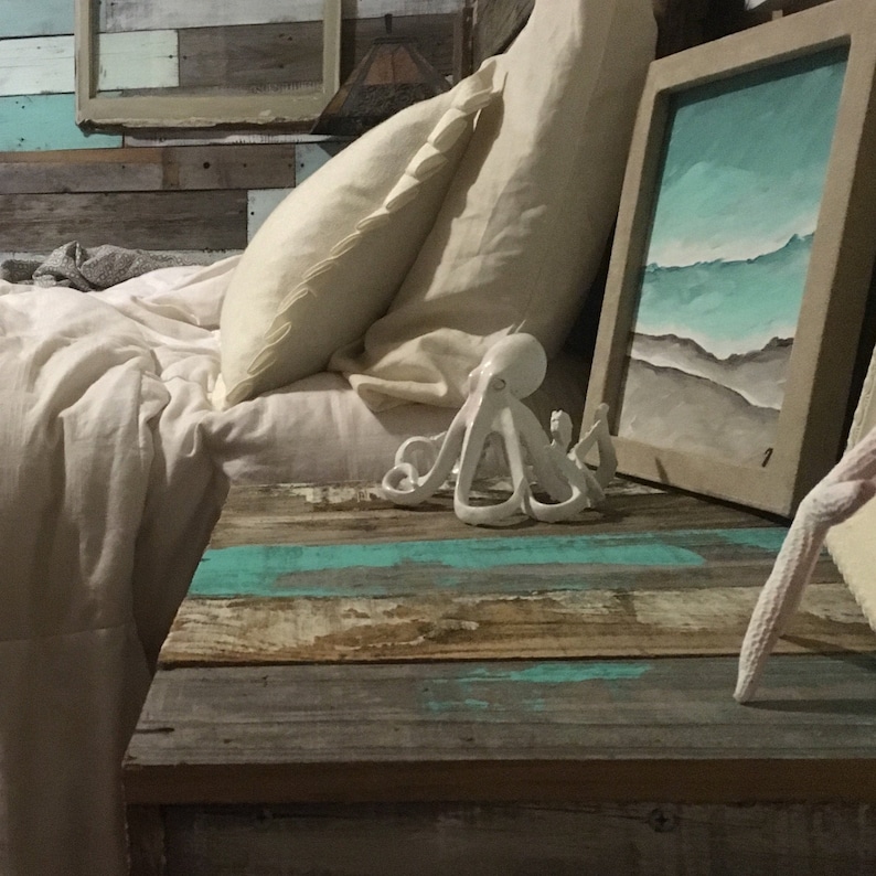 May include: White ceramic octopus figurine on a wooden table with a blue and white painted finish. The table has a white bedspread and pillows behind it. A framed painting with a blue and white seascape is on the table.