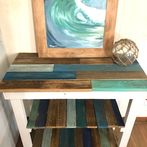 May include: A white wooden table with two shelves. The shelves are made of reclaimed wood planks in shades of blue, green, and brown. A framed painting of a blue ocean wave hangs above the table.