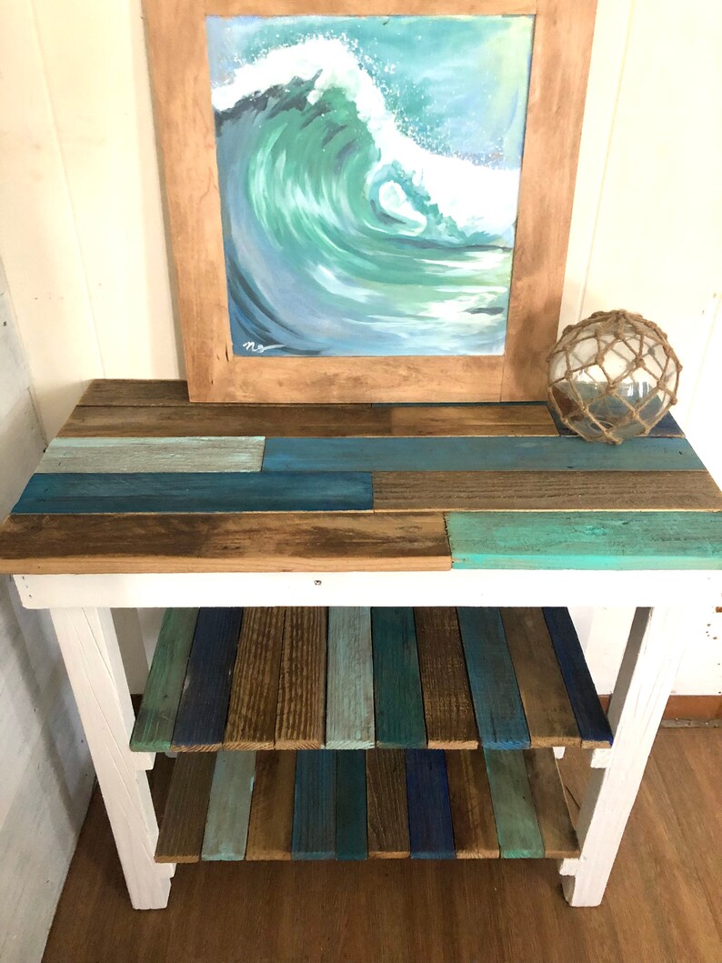 May include: A white wooden table with two shelves. The shelves are made of reclaimed wood planks in shades of blue, green, and brown. A framed painting of a blue ocean wave hangs above the table.