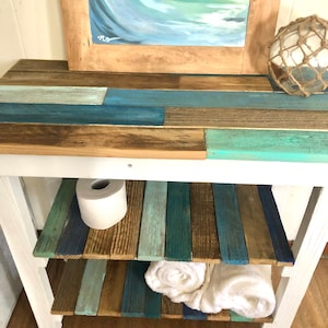 May include: White wooden shelf unit with two shelves. The shelves are made of reclaimed wood planks painted in shades of blue, green, and brown. A roll of toilet paper and two white towels are on the shelves.