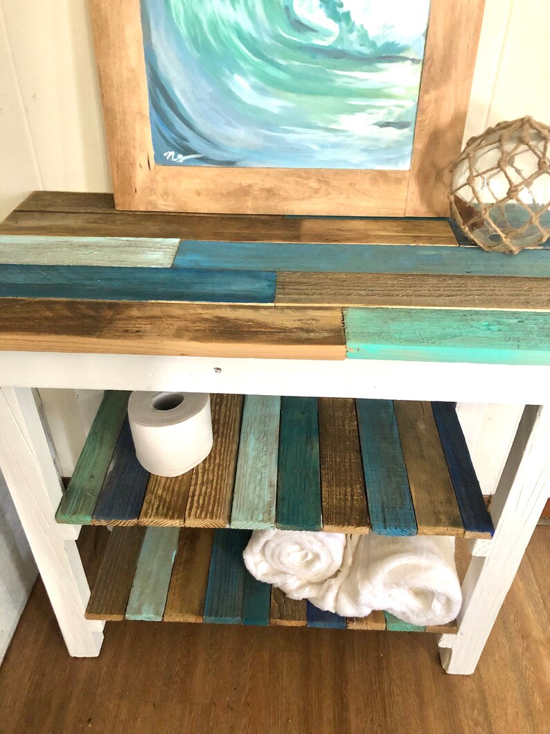 May include: White wooden shelf unit with two shelves. The shelves are made of reclaimed wood planks painted in shades of blue, green, and brown. A roll of toilet paper and two white towels are on the shelves.