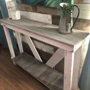 May include: A white painted console table with a gray wood top and a lower shelf. The table has a distressed finish and a simple, farmhouse style. A metal pitcher with a black handle and green plants is on the table.