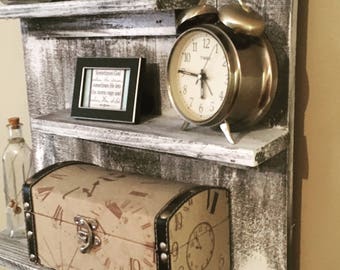 May include: A rustic wooden shelf unit with three shelves, painted white with a distressed finish. The top shelf has a small framed picture with a quote about God, the middle shelf has a vintage alarm clock, and the bottom shelf has a decorative box with a clock face design.